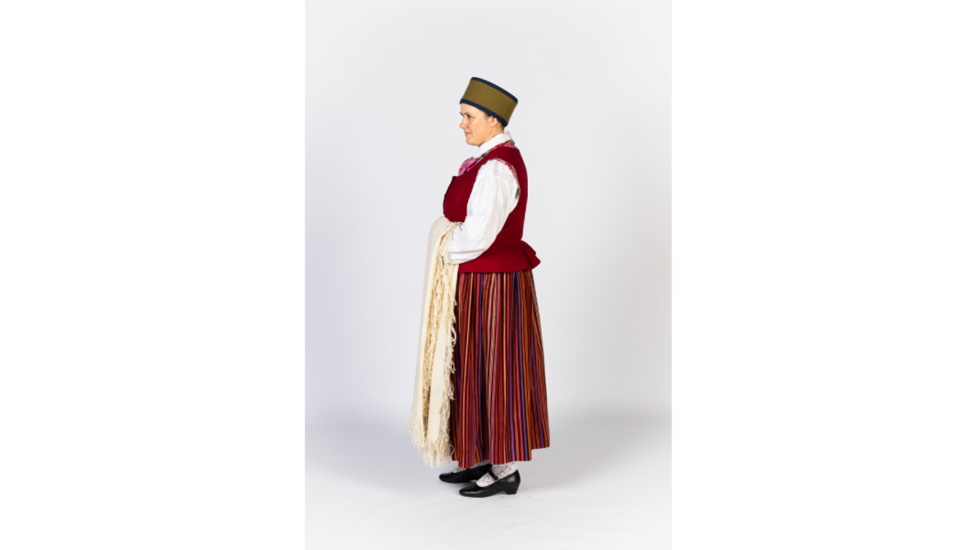 Piebalgas sievas tērps foto no sāniem. Svītraini brunči, keklas, ņieburs, zeķes apavi, galvā ts "zaļmalīte" sievas cepure no melna un zaļa vilnas auduma, rokās austs, balts lakats ar bārkstīm.
