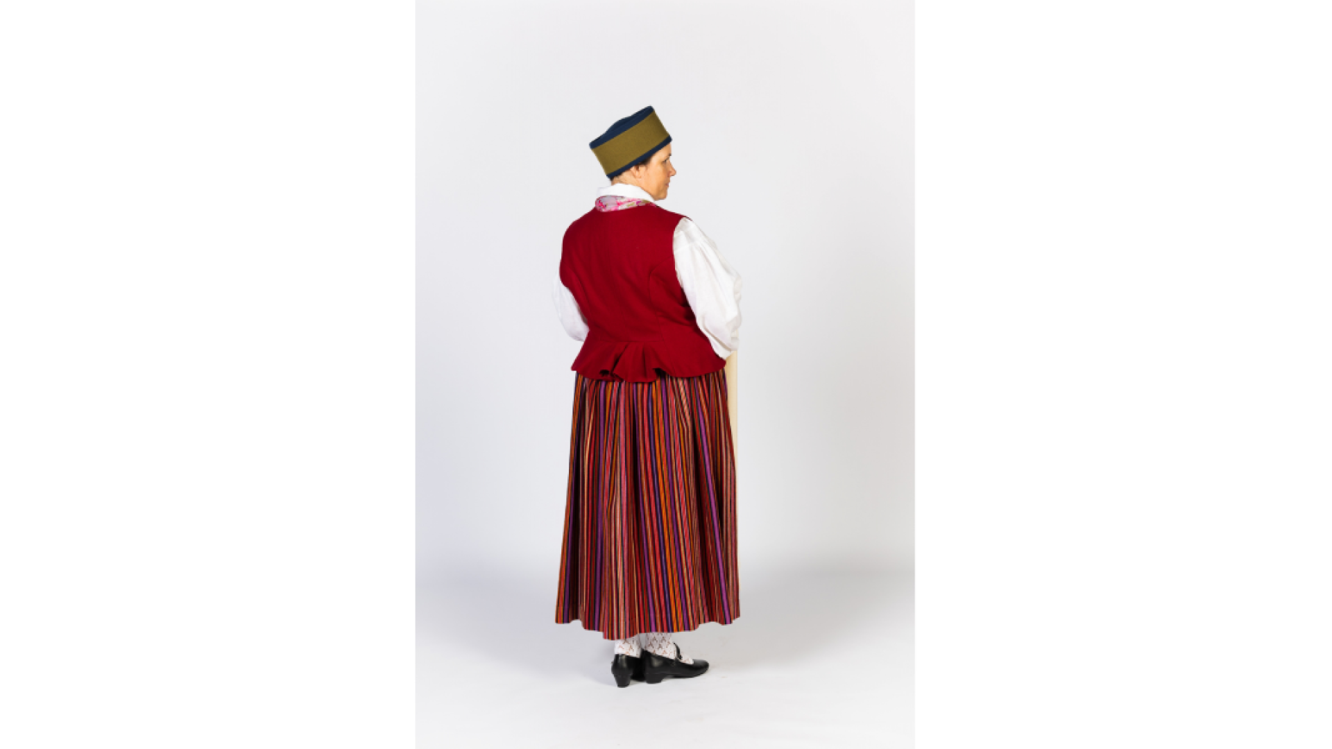Piebalgas sievas tērps foto no mugurpuses. Svītraini brunči, keklas, ņieburs, zeķes apavi, galvā ts "zaļmalīte" sievas cepure no melna un zaļa vilnas auduma, rokās austs, balts lakats ar bārkstīm.