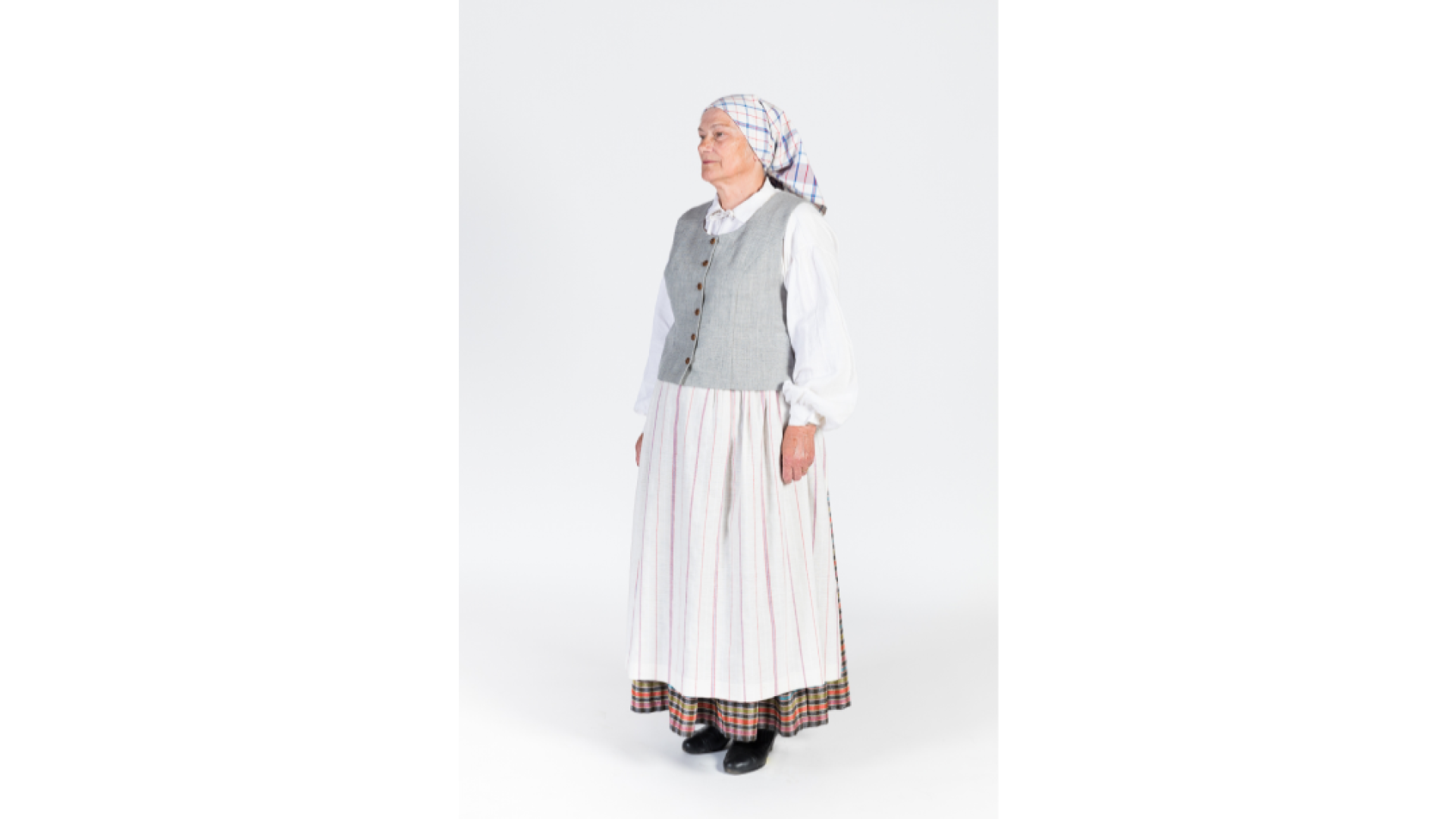Sieva lina un kokvilnas auduma darba tērpā. Balts krekls, rūtaina auduma brunči, pelēka auduma ņieburs, priekšauts, zeļes, apavi, galvā rūtains lakatiņš. Rokās pīta vācele.