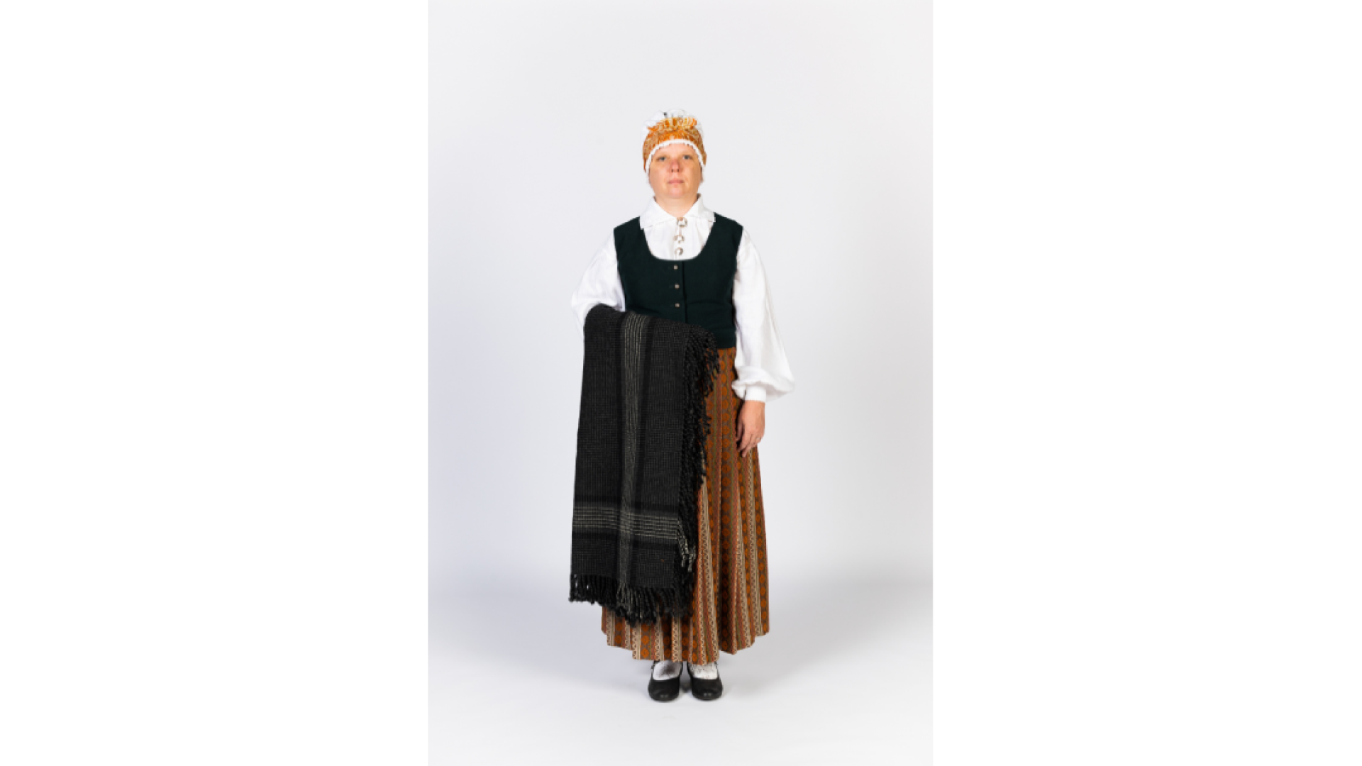 Sieva Zemgales tērpā. Rozīšu brunči, krekls, tumša auduma ņieburs, tilla sievas cepure, kas pārsieta ar lakatiņu, zeķes, kurpes, uz rokas lielais rūtainais lakats pelēkos toņos.